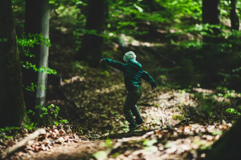 Ein Kind in grüner Jacke springt mit ausgebreiteten Armen über einen Waldweg