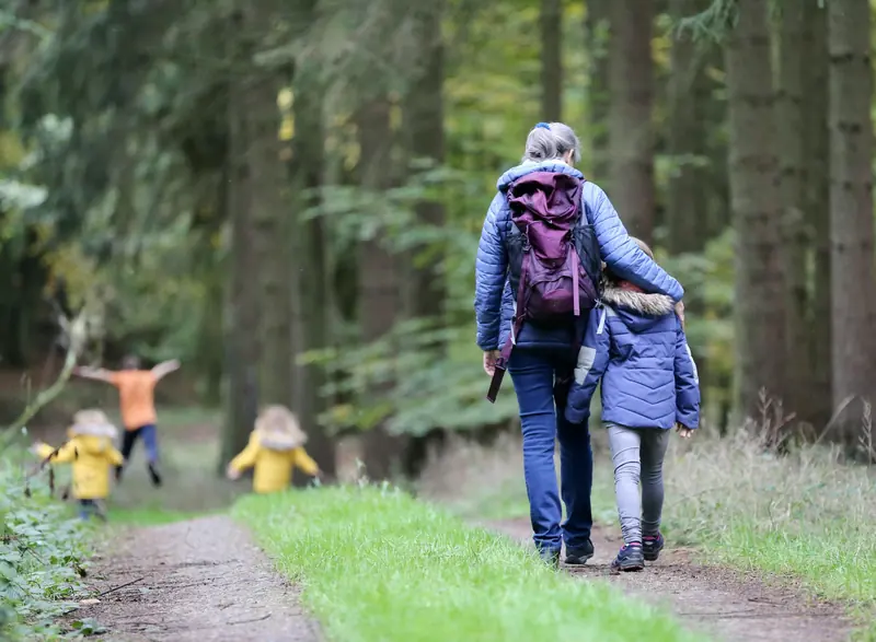 Eine Frau und ein Mädchen gehen Arm in Arm einen Waldweg entlang, dahinter toben zwei Kinder